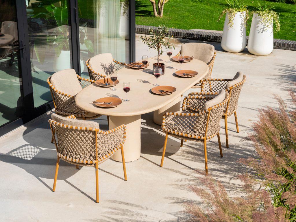 Sechs geflochtene Dining-Stühle aus der Serie Martinique von 4SO um einen ovalen Hampton Tisch mit Tellern, Rotweingläsern, Utensilien und einem Tafelaufsatz. Die Terrasse blickt auf einen grünen Rasen und große weiße Pflanzgefäße neben Glastüren.