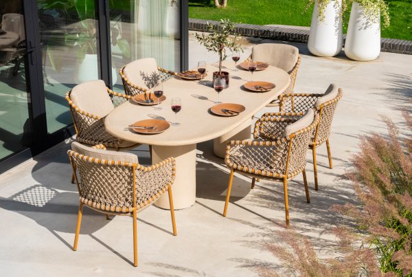Sechs geflochtene Dining-Stühle aus der Serie Martinique von 4SO um einen ovalen Hampton Tisch mit Tellern, Rotweingläsern, Utensilien und einem Tafelaufsatz. Die Terrasse blickt auf einen grünen Rasen und große weiße Pflanzgefäße neben Glastüren.
