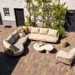 Außenterrasse mit hellbraunem Ziegelsteinboden, gepolstertem Modular-Lounge-Set-Sofa, passendem Living-Sessel aus der Serie Martinique von 4SO, zwei runden weißen Hampton Couchtischen und einem kleinen Beistelltisch aus Korb. Entlang einer weißen Backsteinmauer wachsen Grünpflanzen und Weinreben.