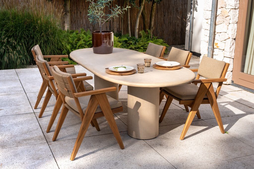 Eine Essgruppe für den Außenbereich aus der Serie Mats von 4SO Gartenmöbel besteht aus einem ovalen Hampton Tisch mit dickem, zylindrischem Fuß, der von sechs hölzernen Dining-Stühlen mit hellbraunen Kissen umgeben ist. Üppiges Grün und Steinmauern vervollständigen die einladende Szene.