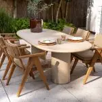 Eine Essgruppe für den Außenbereich aus der Serie Mats von 4SO Gartenmöbel besteht aus einem ovalen Hampton Tisch mit dickem, zylindrischem Fuß, der von sechs hölzernen Dining-Stühlen mit hellbraunen Kissen umgeben ist. Üppiges Grün und Steinmauern vervollständigen die einladende Szene.