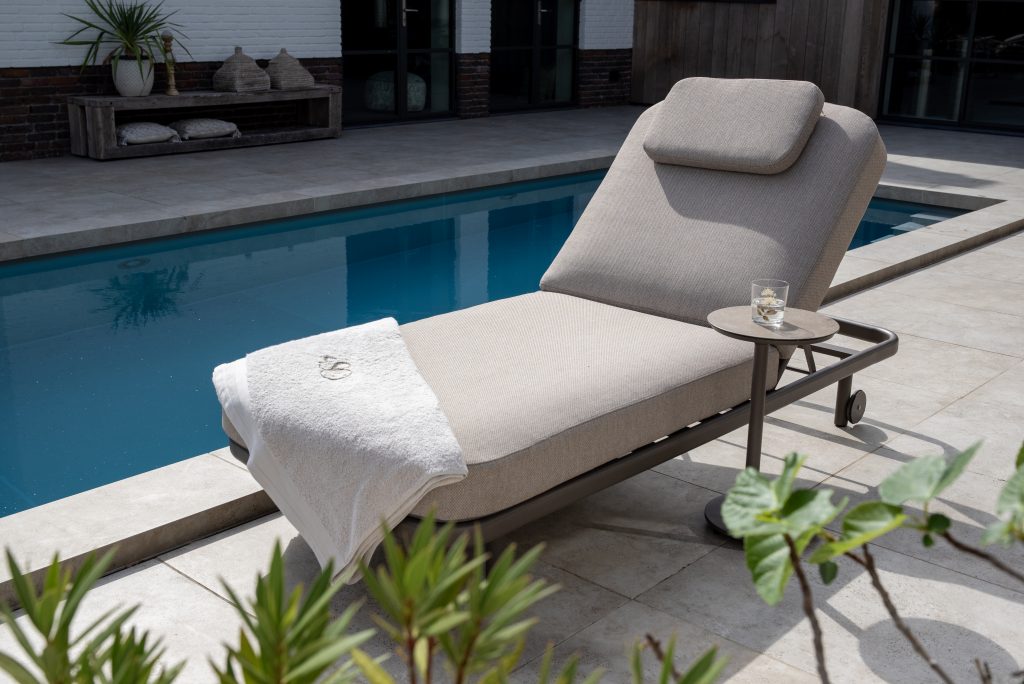 Eine gepolsterte Milano Sonnenliege 200x80 von 4SO in Terre mit Kopfstütze steht an einem blau gekachelten Pool. Ein weißes Handtuch ist über das Ende drapiert, und auf einem kleinen runden Tisch steht ein Glas Wasser. Üppige Grünpflanzen und Gartenmöbel vervollständigen die Szene.