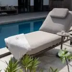 Eine gepolsterte Milano Sonnenliege 200x80 von 4SO in Terre mit Kopfstütze steht an einem blau gekachelten Pool. Ein weißes Handtuch ist über das Ende drapiert, und auf einem kleinen runden Tisch steht ein Glas Wasser. Üppige Grünpflanzen und Gartenmöbel vervollständigen die Szene.