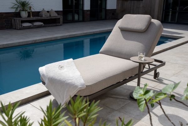 Eine gepolsterte Milano Sonnenliege 200x80 von 4SO in Terre mit Kopfstütze steht an einem blau gekachelten Pool. Ein weißes Handtuch ist über das Ende drapiert, und auf einem kleinen runden Tisch steht ein Glas Wasser. Üppige Grünpflanzen und Gartenmöbel vervollständigen die Szene.