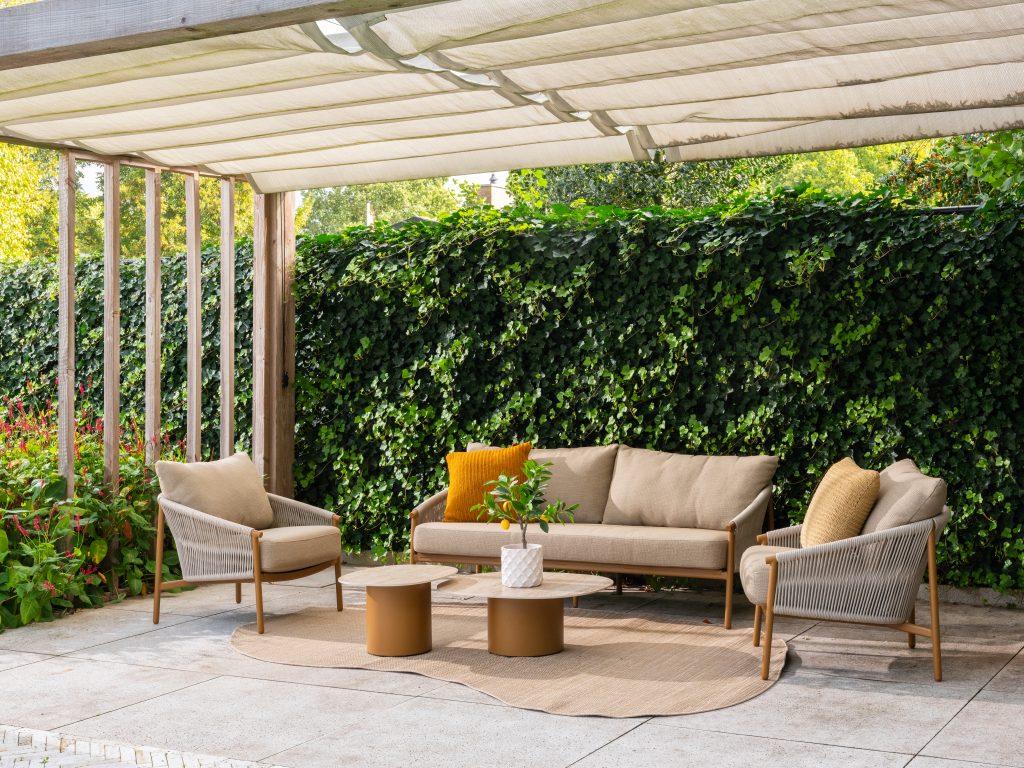 Ein moderner Freisitz unter einer Pergola mit Gartenmöbeln aus der Serie Milos von 4SO: ein bernsteinfarbenes Living-Sofa und zwei Sessel, ein runder Teppich, zwei niedrige Sofie Tische und Deko-Kissen. Üppig grüner Efeu bedeckt den Zaun mit Pflanzen an den Seiten.