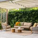 Ein moderner Freisitz unter einer Pergola mit Gartenmöbeln aus der Serie Milos von 4SO: ein bernsteinfarbenes Living-Sofa und zwei Sessel, ein runder Teppich, zwei niedrige Sofie Tische und Deko-Kissen. Üppig grüner Efeu bedeckt den Zaun mit Pflanzen an den Seiten.