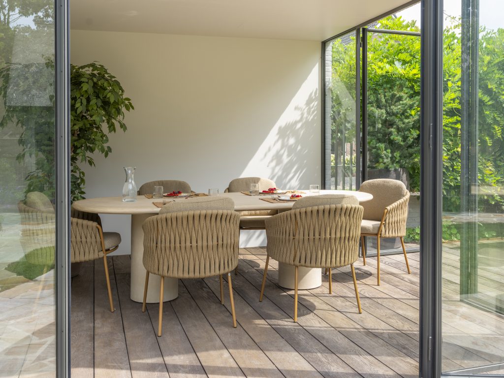In einem modernen Esszimmer mit Glaswänden steht ein Hampton Tisch aus hellem Holz, umgeben von sechs Dining-Stühlen der Serie Molina von 4SO auf einem Holzboden. Eine große Blattpflanze sorgt für Frische, und durch die Fenster ist üppiges Laub im Freien zu sehen.