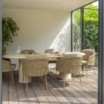 In einem modernen Esszimmer mit Glaswänden steht ein Hampton Tisch aus hellem Holz, umgeben von sechs Dining-Stühlen der Serie Molina von 4SO auf einem Holzboden. Eine große Blattpflanze sorgt für Frische, und durch die Fenster ist üppiges Laub im Freien zu sehen.