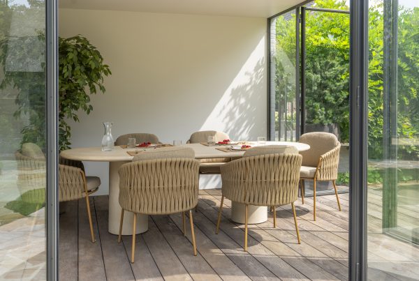 In einem modernen Esszimmer mit Glaswänden steht ein Hampton Tisch aus hellem Holz, umgeben von sechs Dining-Stühlen der Serie Molina von 4SO auf einem Holzboden. Eine große Blattpflanze sorgt für Frische, und durch die Fenster ist üppiges Laub im Freien zu sehen.