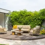 Eine moderne Terrasse mit Living-Sofa und Sessel der Serie Molina von 4SO, davor zwei runde Sofie Tische mit Pflanzen. Die ovale Holzterrasse wird von einem Steinweg, üppigen grünen Sträuchern und gelben Blumen gesäumt. Entdecken Sie Gartenmöbel bei 1001-Sommer.