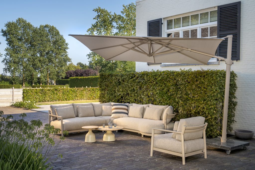Eine moderne Außenterrasse mit einem großen Modular-Lounge-Sofa, einem passenden Living-Sessel und zwei runden June Couchtischen. Ein großer, freitragender Sonnenschirm spendet Schatten. Entdecken Sie Gartenmöbel bei 1001-Sommer kaufen für Stil wie die Serie Mykonos von 4SO.