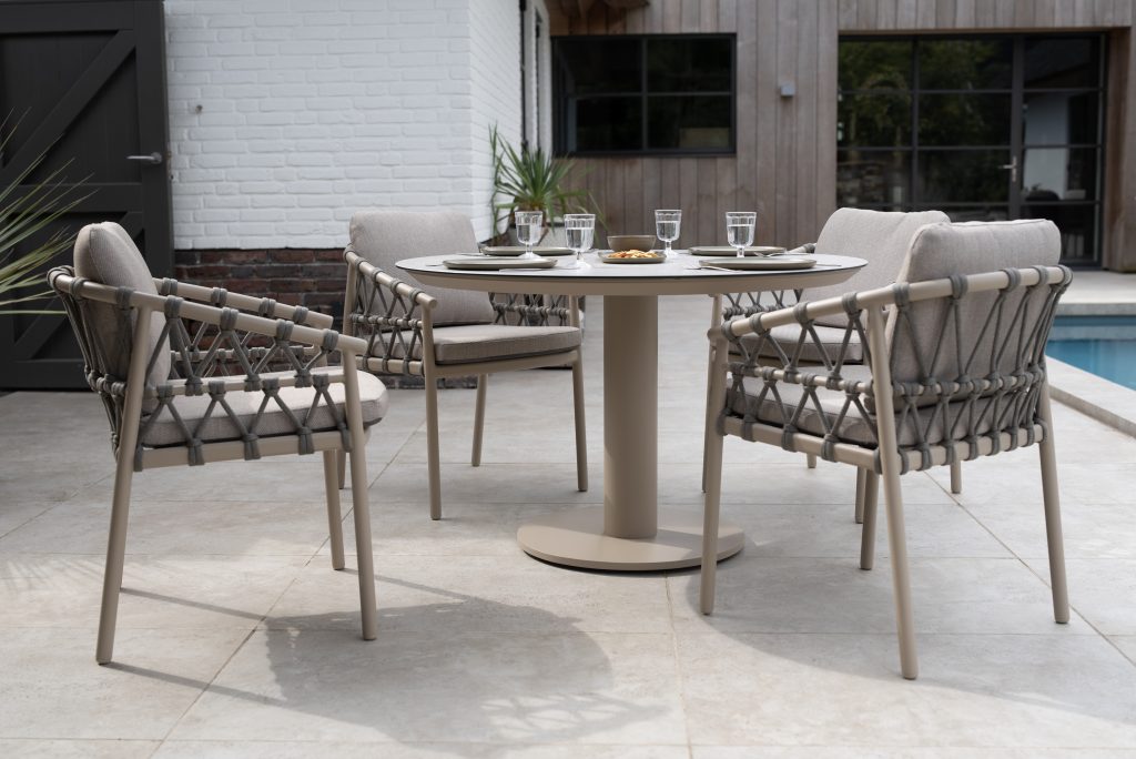 Ein modernes Terrassenset mit der Serie Piacenza von 4SO Gartenmöbel mit vier gepolsterten Dining-Stühlen um einen runden Sarah-Tisch mit Tellern, Gläsern und einer Schale auf einer gefliesten Terrasse am Pool, umrahmt von Pflanzen und einer weißen Backsteinmauer.