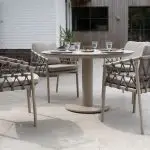 Ein modernes Terrassenset mit der Serie Piacenza von 4SO Gartenmöbel mit vier gepolsterten Dining-Stühlen um einen runden Sarah-Tisch mit Tellern, Gläsern und einer Schale auf einer gefliesten Terrasse am Pool, umrahmt von Pflanzen und einer weißen Backsteinmauer.
