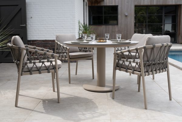 Ein modernes Terrassenset mit der Serie Piacenza von 4SO Gartenmöbel mit vier gepolsterten Dining-Stühlen um einen runden Sarah-Tisch mit Tellern, Gläsern und einer Schale auf einer gefliesten Terrasse am Pool, umrahmt von Pflanzen und einer weißen Backsteinmauer.