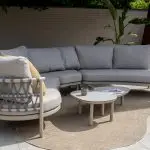 Ein modernes Modular-Lounge-Set Piacenza von 4SO für den Außenbereich besteht aus einem geschwungenen grauen Gartenmöbel-Sectional-Sofa mit gewebten Details, hellen Kissen und einem gelb gestreiften Kopfkissen. Zwei verschachtelte Monroe Couchtische stehen auf einem beigen Teppich, mit Pflanzen und einem hohen braunen Außenkamin im Hintergrund.