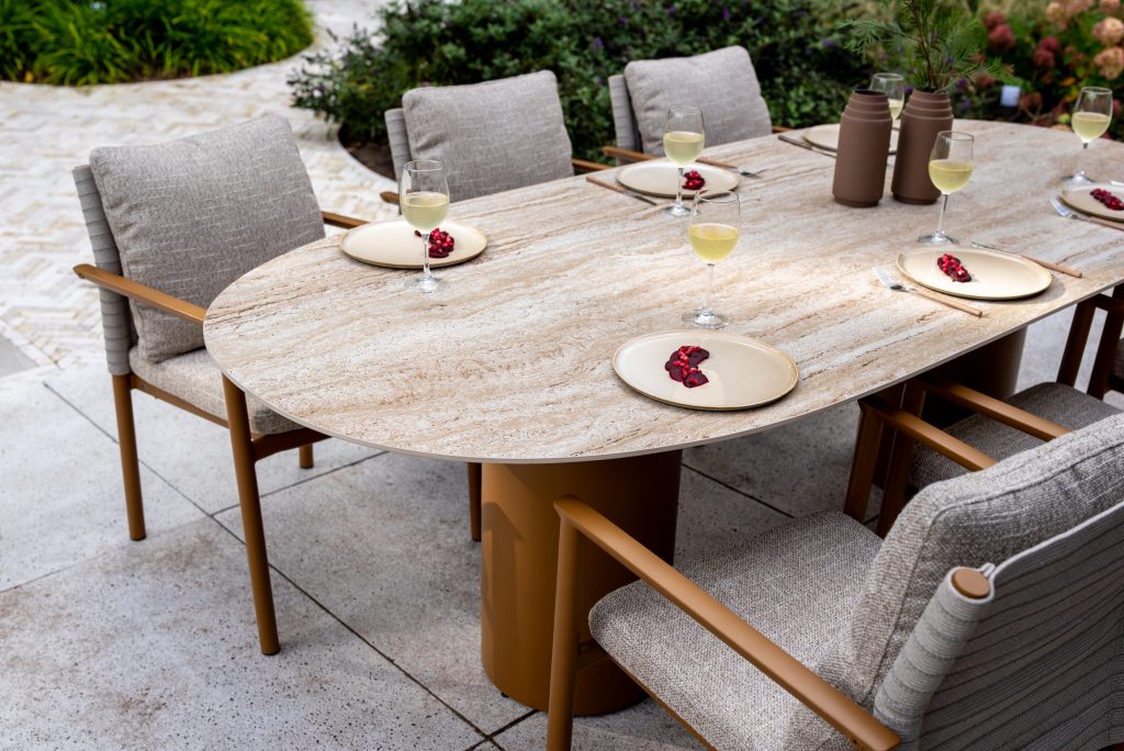 Ein moderner Colorado Diningtisch Oval mit Keramik/Travertin Oberfläche und tischbein bernsteinfarben, umgeben von sechs gepolsterten Stühlen. Auf jedem beigen Teller liegen Granatapfelkerne neben einem Glas Weißwein. Dekorative Vasen akzentuieren die üppige Bepflanzung der Steinterrasse.
