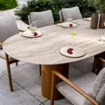 Ein moderner Colorado Diningtisch Oval mit Keramik/Travertin Oberfläche und tischbein bernsteinfarben, umgeben von sechs gepolsterten Stühlen. Auf jedem beigen Teller liegen Granatapfelkerne neben einem Glas Weißwein. Dekorative Vasen akzentuieren die üppige Bepflanzung der Steinterrasse.