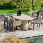 Auf einer Außenterrasse steht ein ovaler Hampton Tisch, umgeben von acht Dining-Stühlen der Serie Savanne von 4SO, die jeweils mit hellen Kissen ausgestattet sind. Schwarze Vasen mit getrockneten Gräsern schmücken den Tisch, der von Ziergras, Backsteinmauern und einem üppigen grünen Rasen umgeben ist.