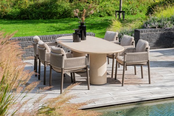 Auf einer Außenterrasse steht ein ovaler Hampton Tisch, umgeben von acht Dining-Stühlen der Serie Savanne von 4SO, die jeweils mit hellen Kissen ausgestattet sind. Schwarze Vasen mit getrockneten Gräsern schmücken den Tisch, der von Ziergras, Backsteinmauern und einem üppigen grünen Rasen umgeben ist.