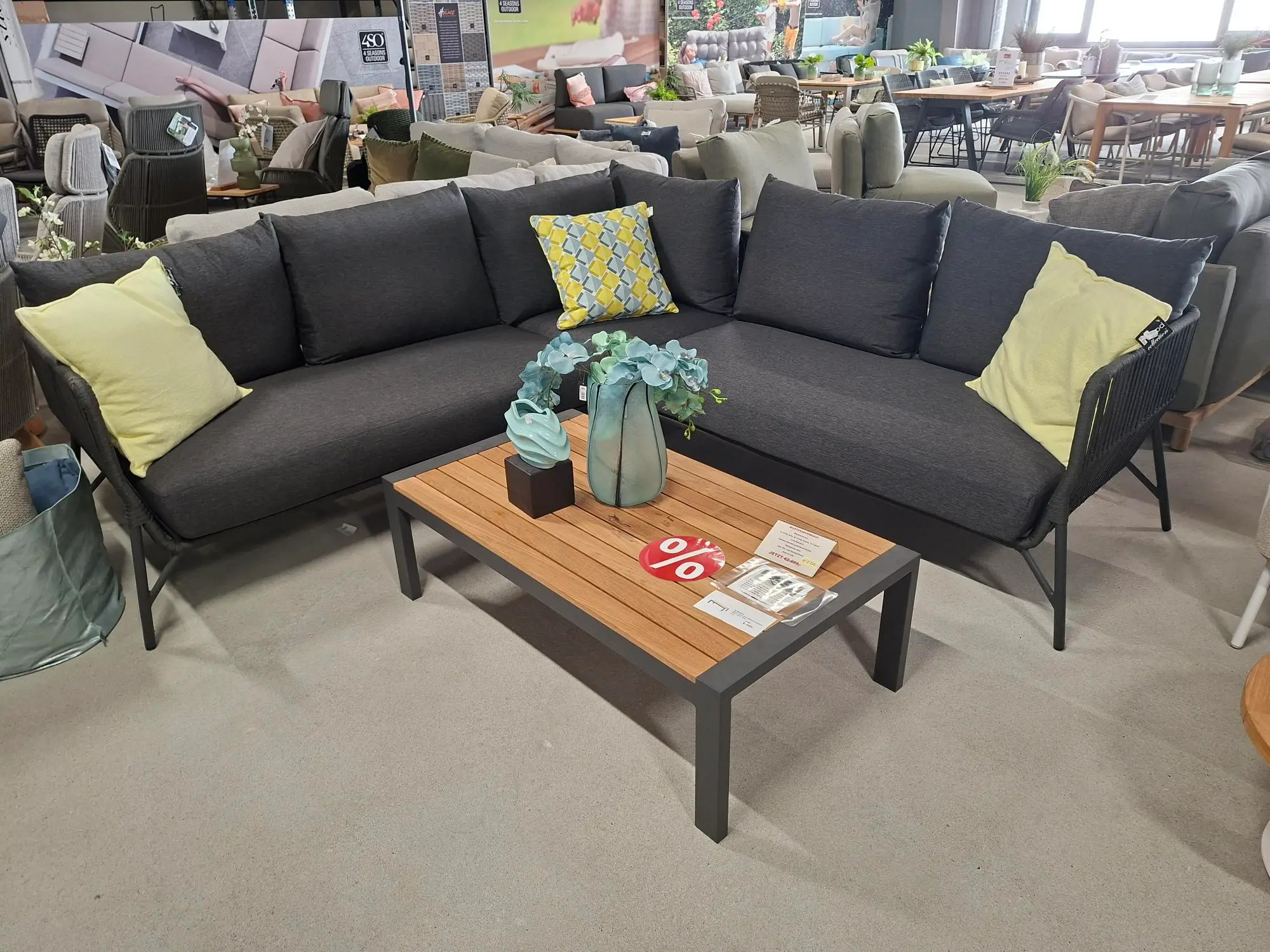 Hinter einem Couchtisch aus Holz steht ein modernes, L-förmiges, dunkelgraues Sofa mit hellgelben und gemusterten Kissen. Auf dem Tisch stehen eine Vase mit blauen Blumen, ein Prozentzeichen, das auf einen Sonderverkauf hinweist, sowie zwei Broschüren. Der Schauplatz ist ein Möbelausstellungsraum mit verschiedenen Ausstellungsstücken.